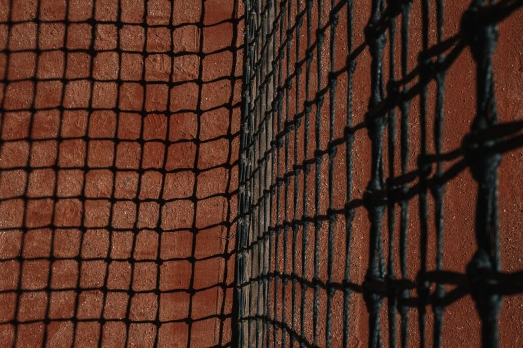 Detailed close-up of a tennis net casting shadows on a clay court surface during the day.